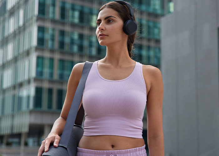Young woman wearing headphones and gym clothes, carrying a yoga mat in an urban setting, reflecting on men’s comments about looks.