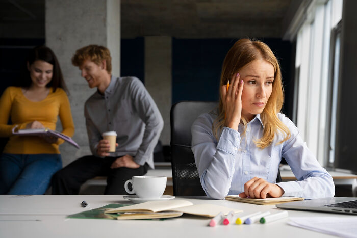 Woman looking distressed at desk while two colleagues in background chat and laugh, depicting signs of childhood struggles