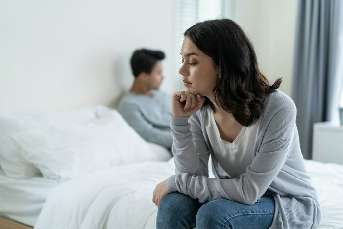A woman looking thoughtful and distant while her partner sits on the bed, representing secrets people keep from their lovers.