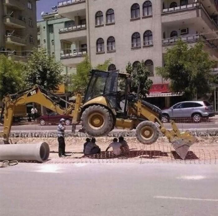Men doing dumb things sitting under lifted heavy construction equipment on a city street during daytime.
