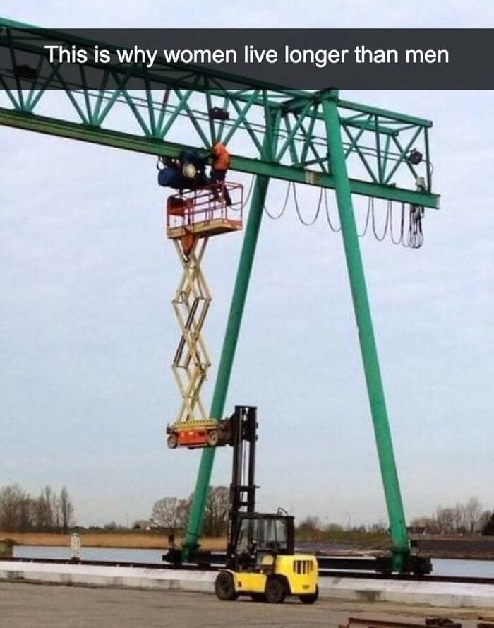 Man doing dumb things by standing on a scissor lift raised by a forklift, illustrating why women live longer.